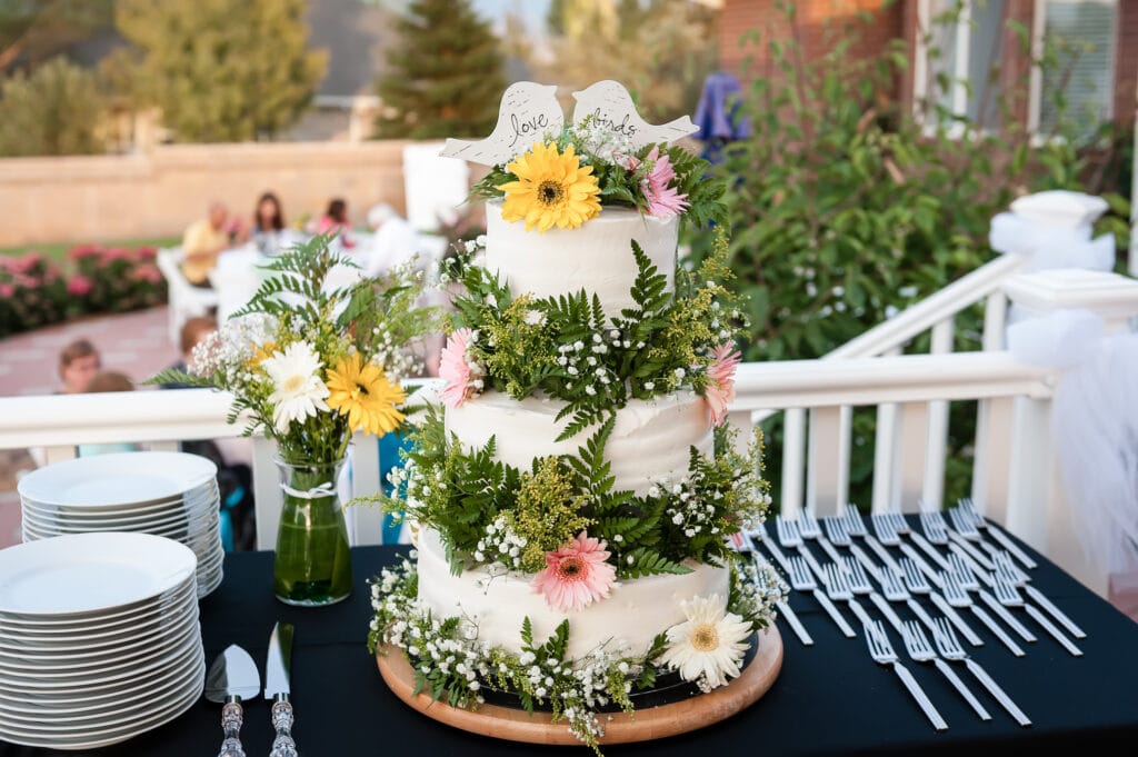 A three-tiered hand-crafted wedding cake decorated with greenery and Gerbera daisies and two cut-outs of love birds on top.