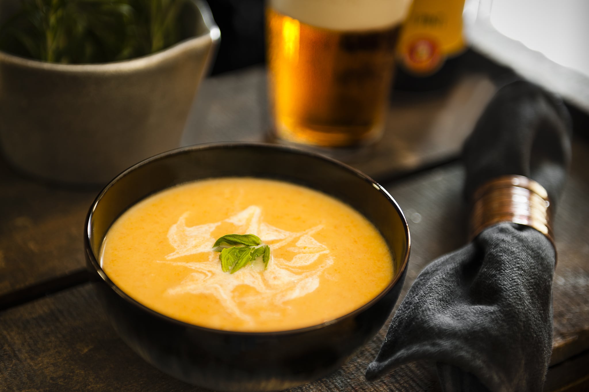 A bowl of soup with a glass of beer in the background.