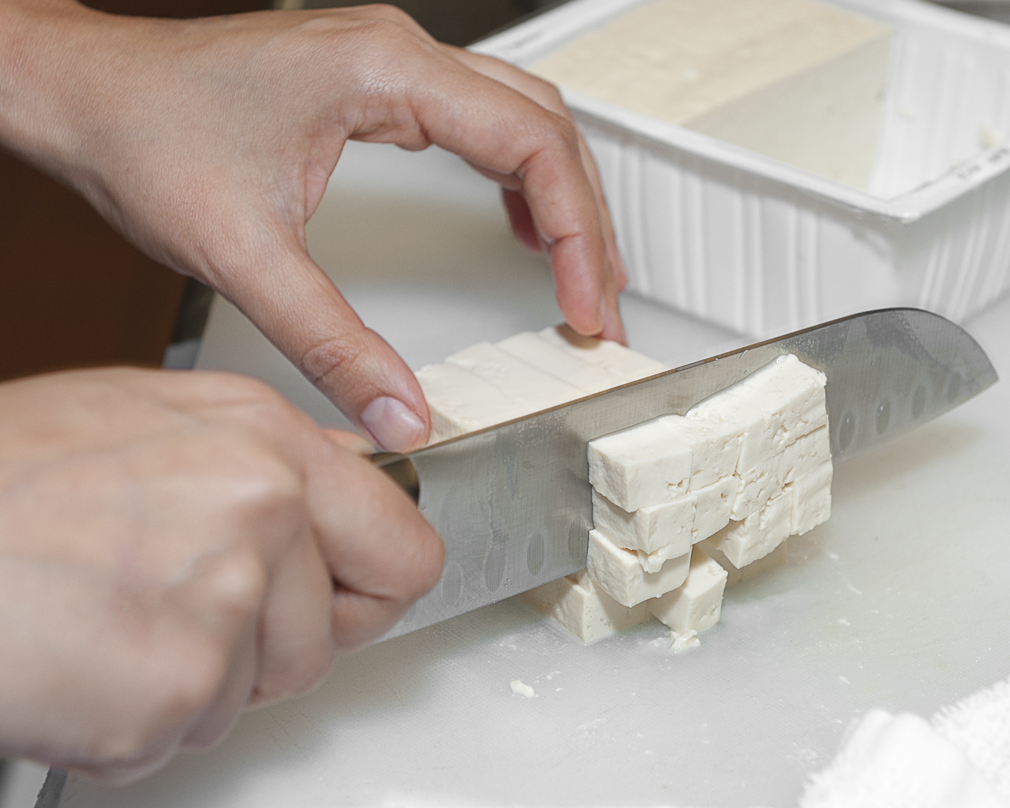 Close up of the hands of a woman as she cuts a block of tofu with a chef knife into small cubes.