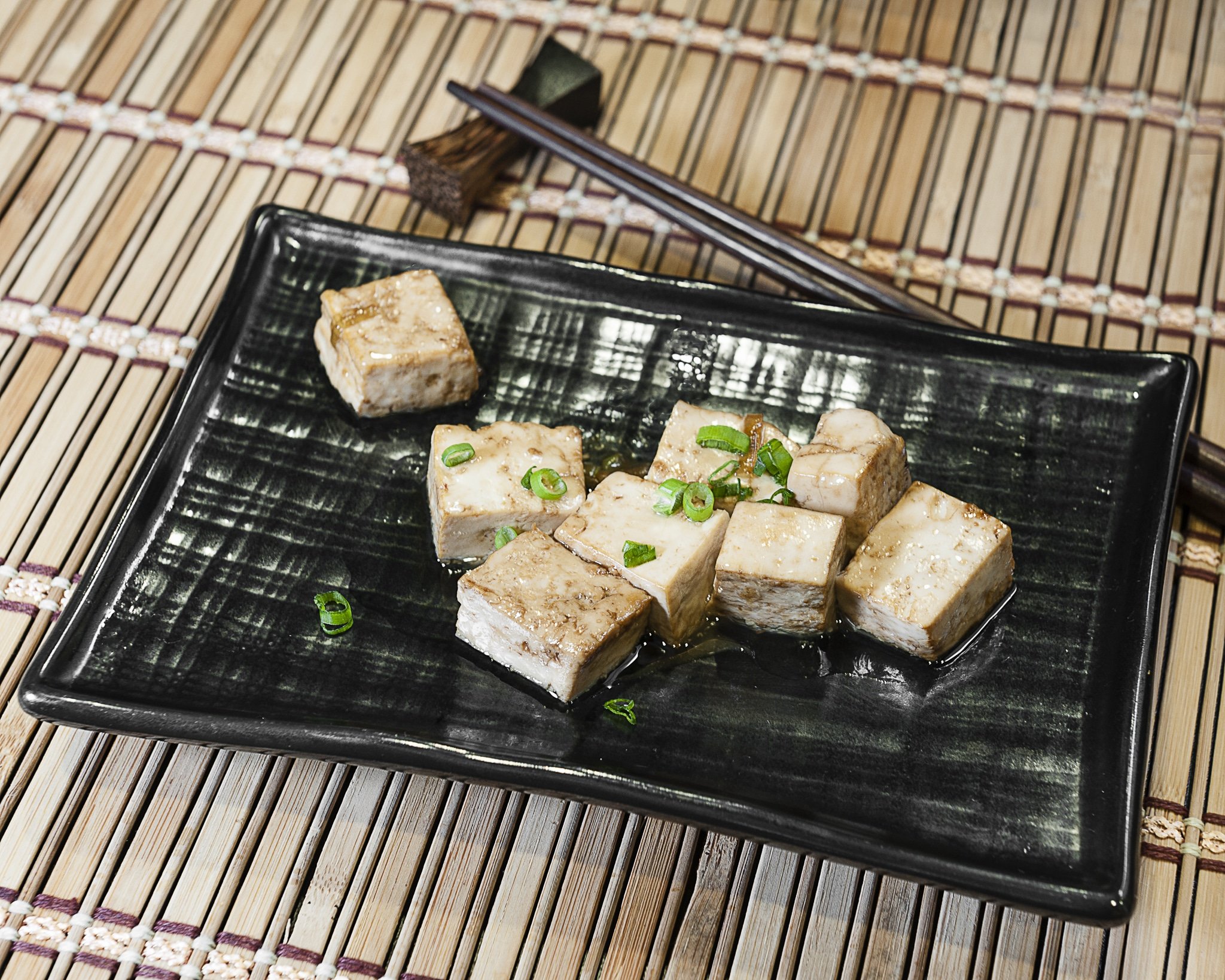 Prepared tofu dish with scallions on a black rectangular serving tray sitting on top of a bamboo mat.