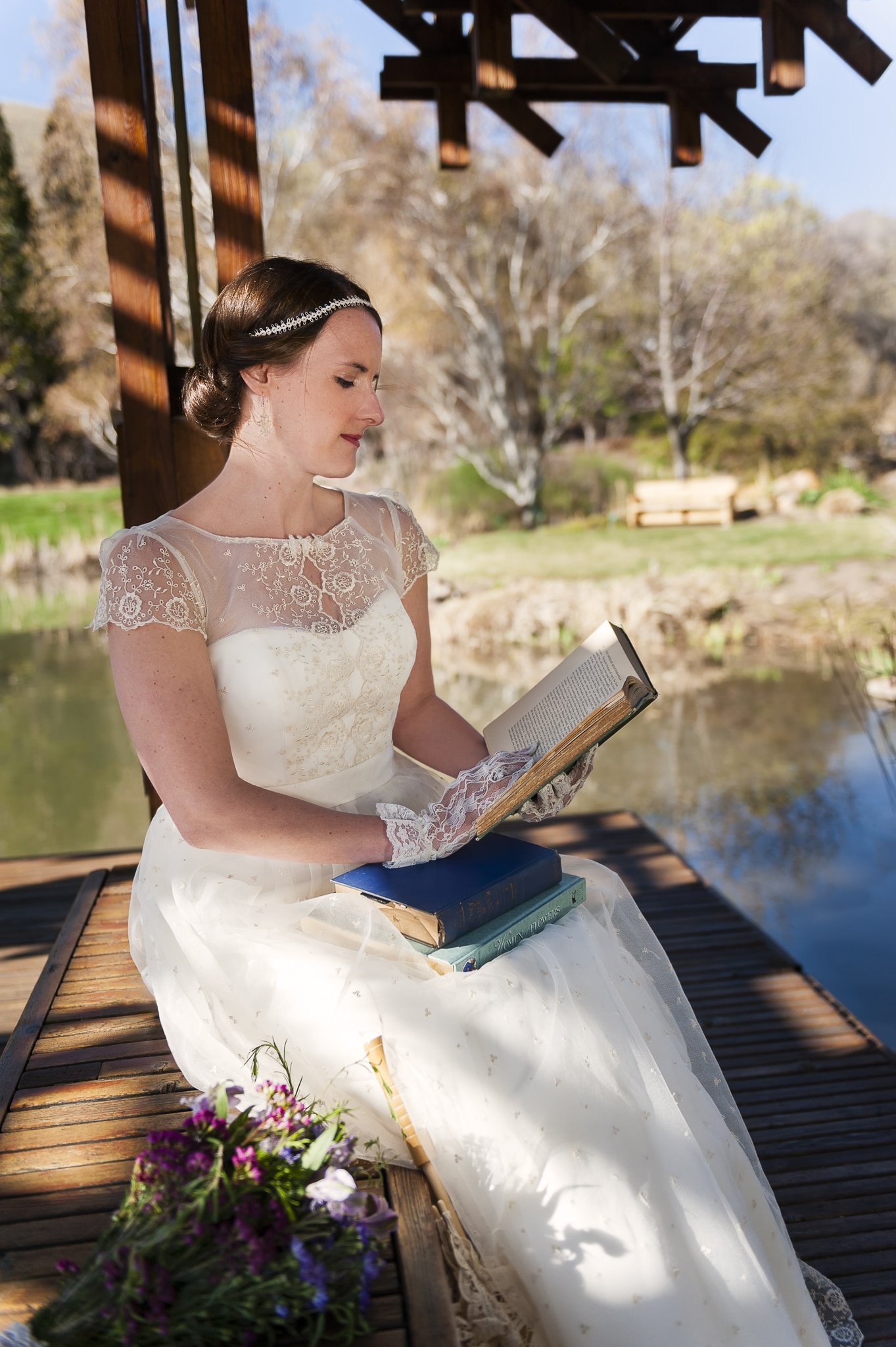 A bride-to-be in wedding attire reads some of her favorite books under a shaded gazebo during her bridal portrait session in a garden.