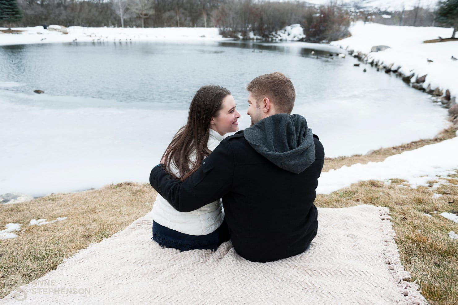 A man puts his arm around his fiance as they sit on a blanket together and watch ducks in a partially frozen pond during their engagement session.