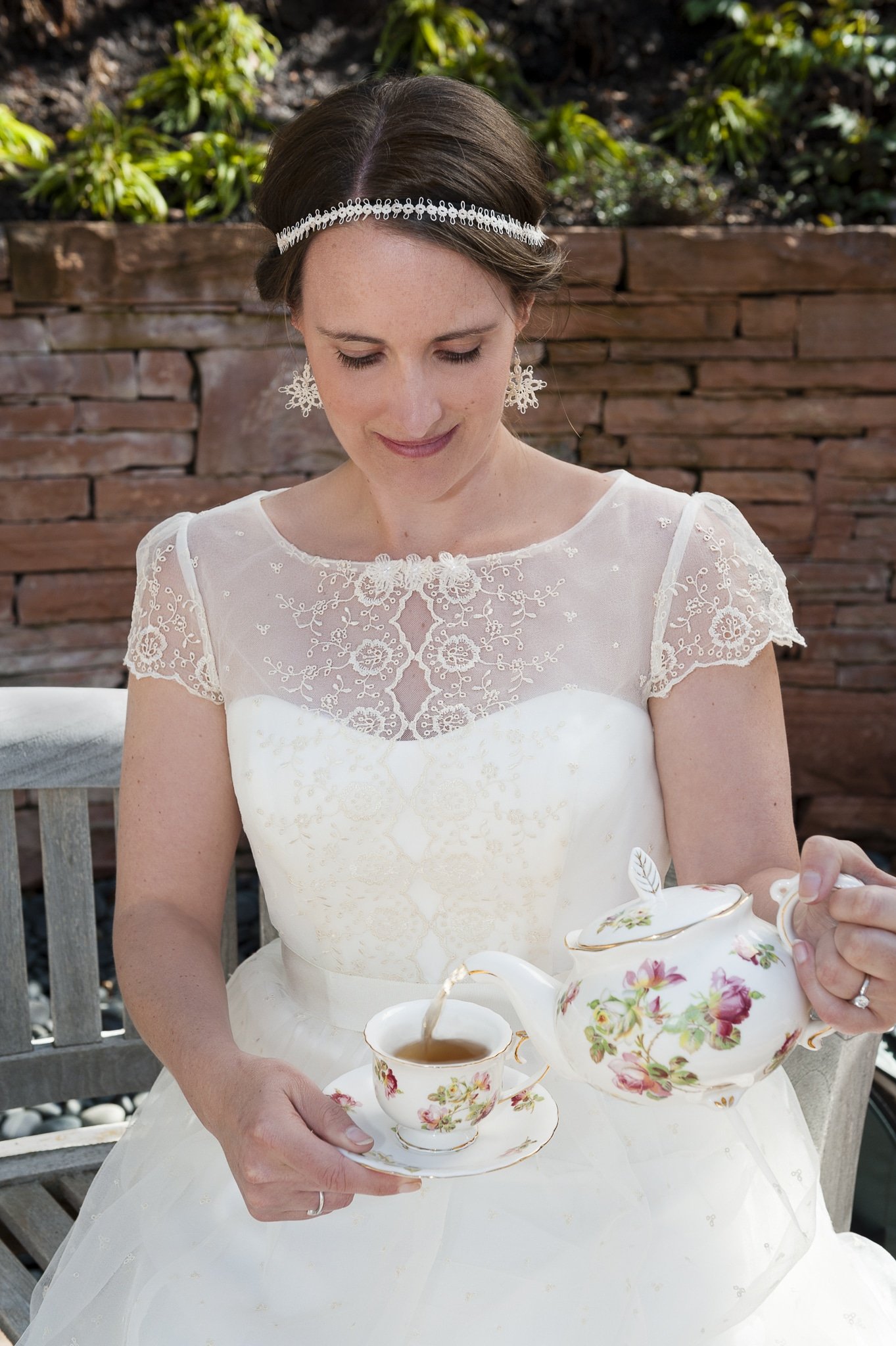 A bride-to-be in wedding attire pours tea from a traditional teapot during her bridal portrait session in a garden.