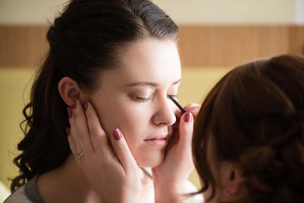 A bride receives help from the artistry of her friend in applying her makeup.