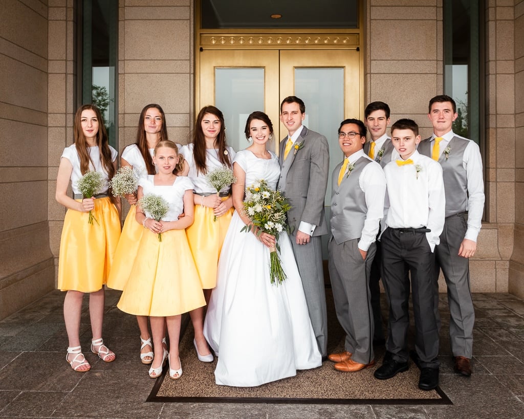 Wedding party in yellow and gray stand in front of the temple's gold doors underneath the protection of the temple alcove.