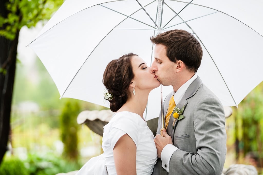 Newlyweds share a kiss together in the rain under the protection of a white umbrella.