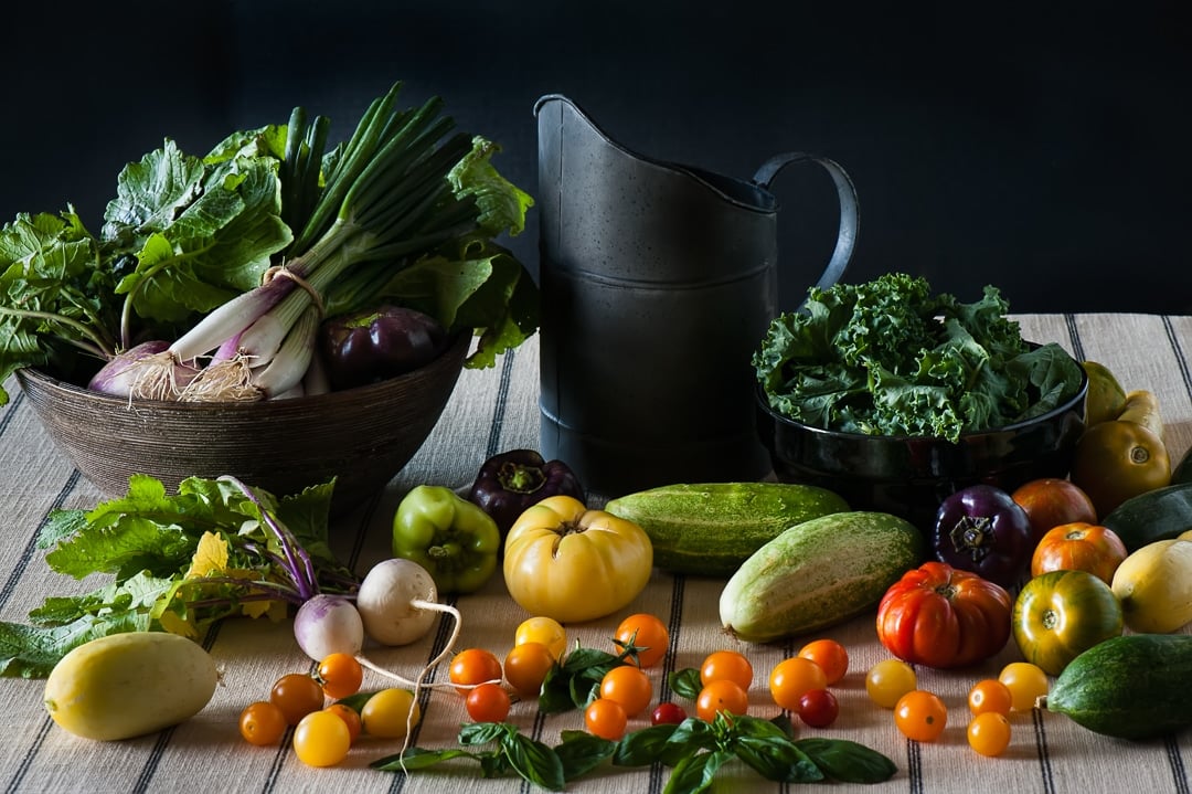 A still life food scene of a bounty of fresh produce including tomato, cucumber, basil, onion, and kale along with a dark black metal pitcher.