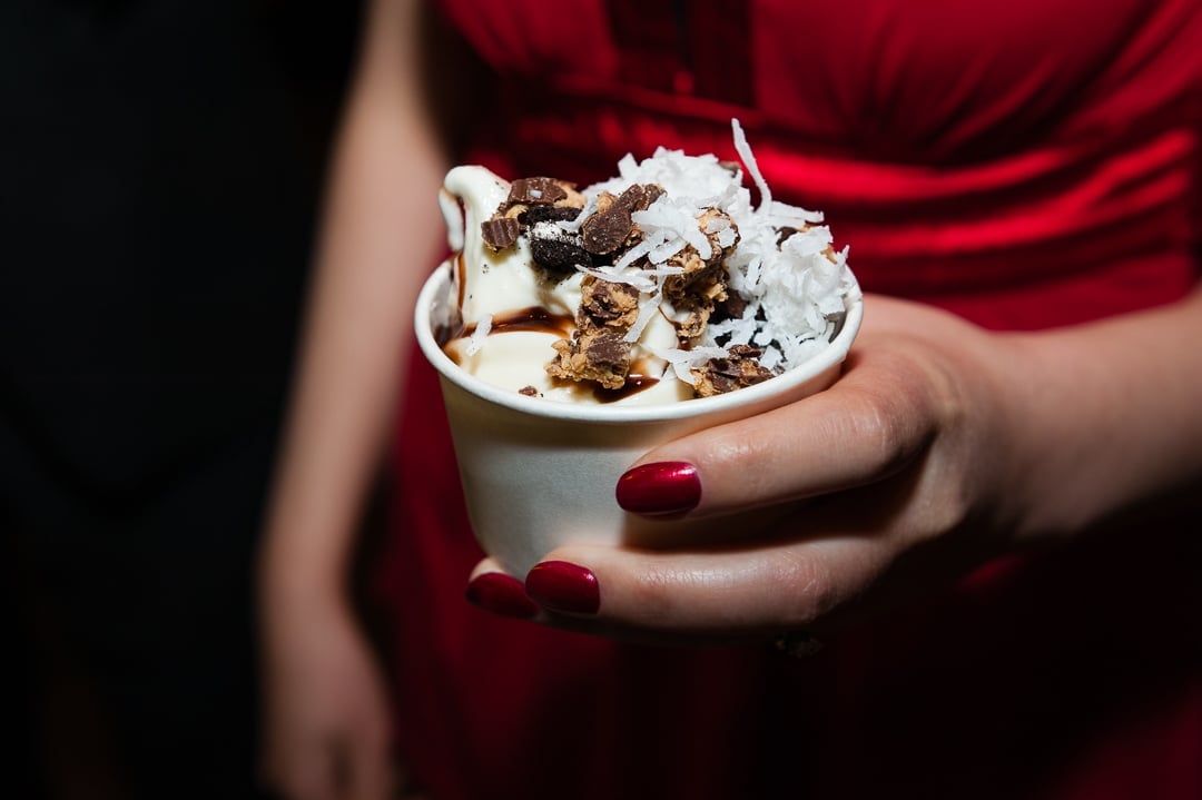 A woman wearing bright right and matching red nail polish holds a container with a scoop of ice cream topped with crunch candy bar, caramel, and coconut.