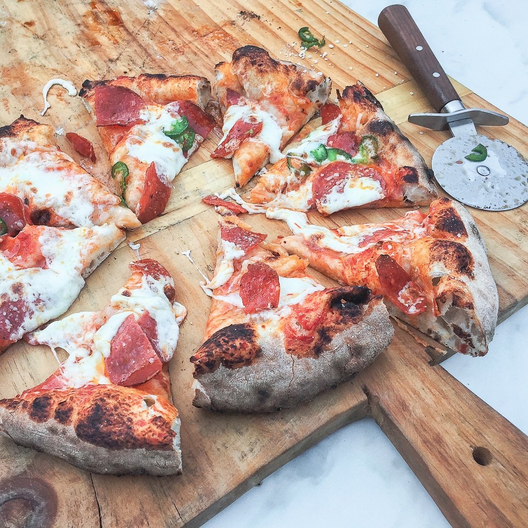 A wood-fired pizza with pepperoni and vegetables sliced into pieces and pizza slicer.