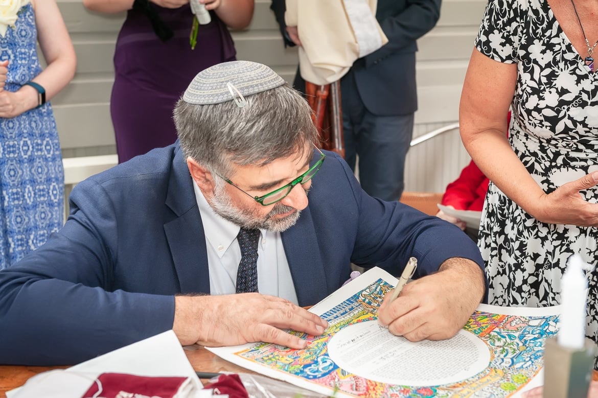 A rabbi kneels down to sign the ketubah at an outdoor Jewish ceremony.