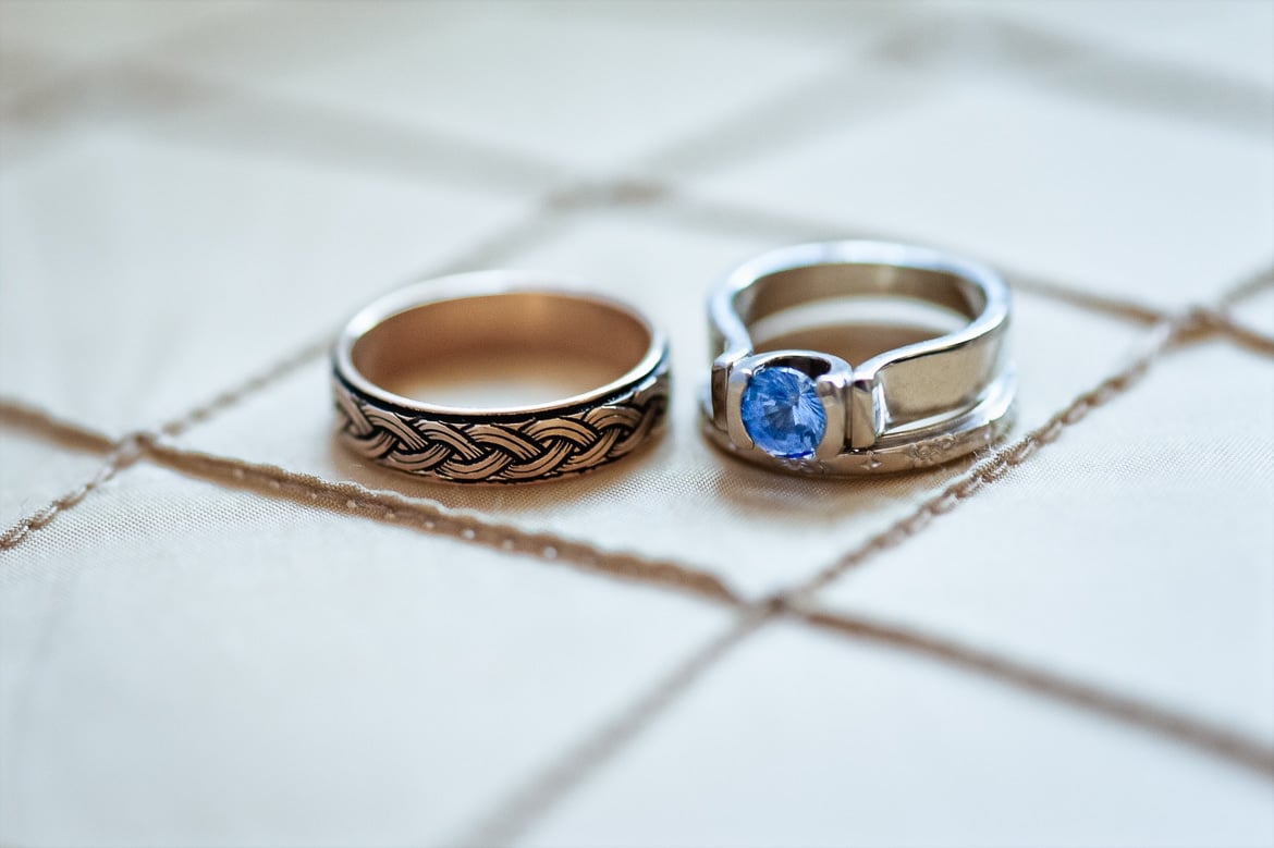 Close up of two wedding rings, one with etching and the other with a pretty blue stone, on a light-colored tan quilt.