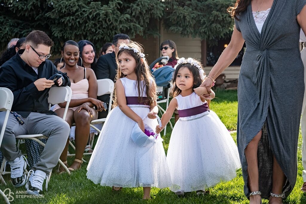 Two flower girls in white flowing dresses and purple sashes walk down the aisle together.