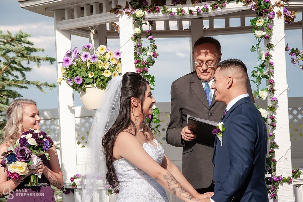 The bride and groom under the white gazebo hold hands and exchange vows in front of the officiant.