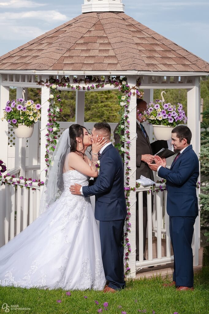 The bride and groom share a first kiss under the flower-strewn gazebo as the best man applauds.