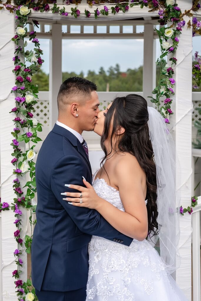 The recently married bride and groom share a passionate kiss under the gazebo strewn with purple flowers at Crystal Rose.
