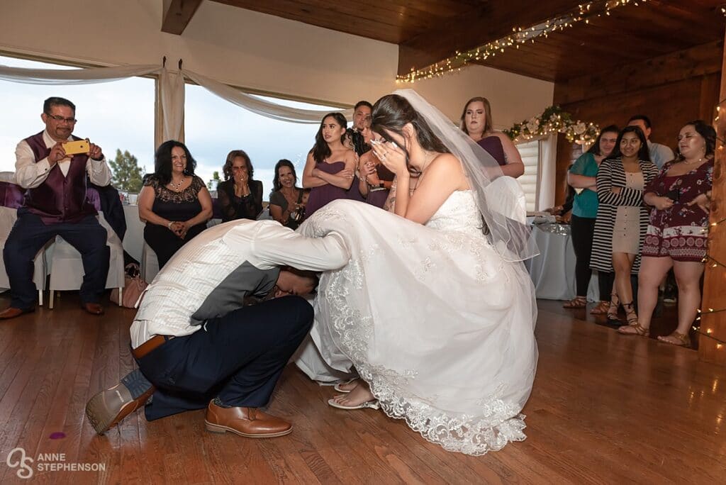 The blushing bride covers her face as the groom retrieves the garter from her leg.