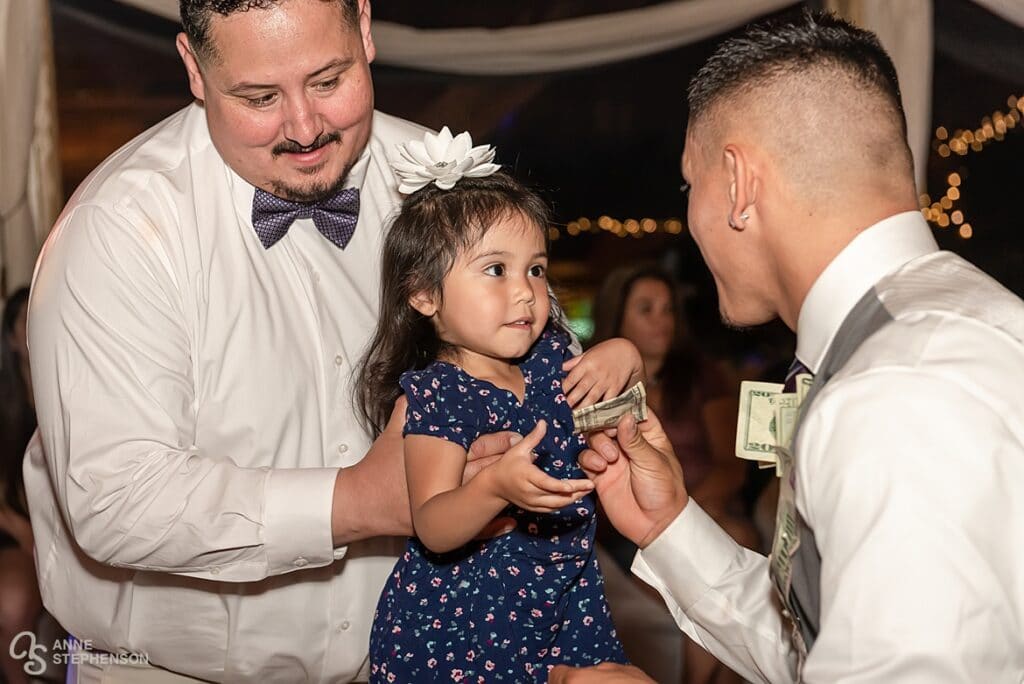 A young girl held by her father gives the groom dollar bills during the traditional wedding money dance.