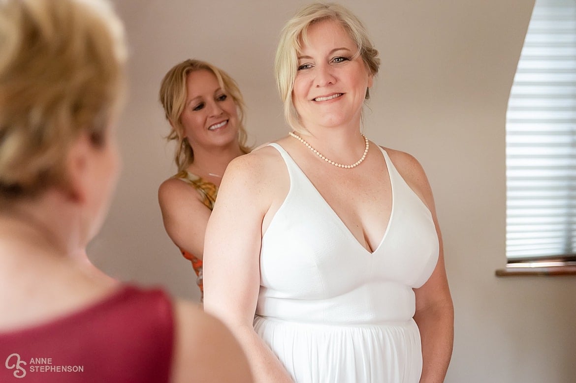 A bride smiles as her maid of honor helps with the dress as the mother of the bride looks on.