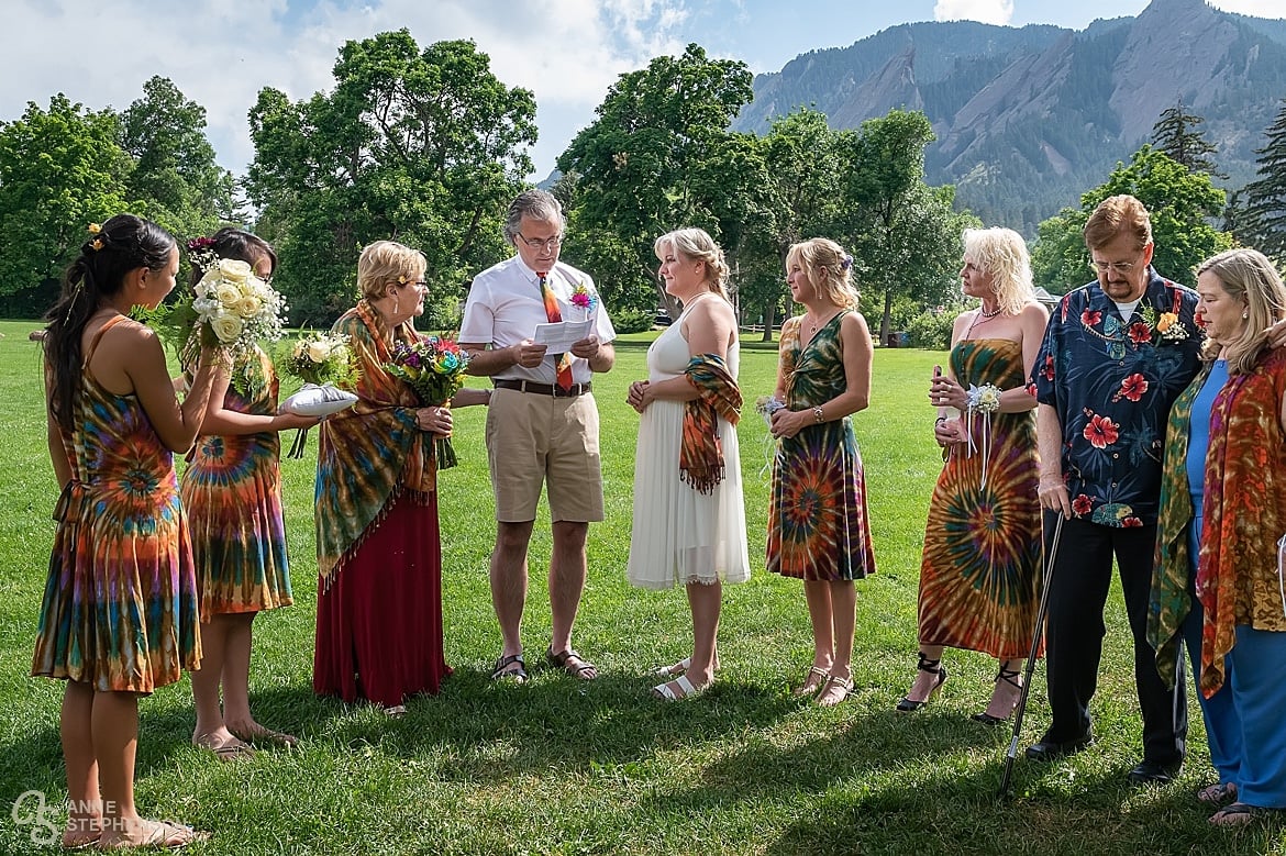 A half circle of friends and family in tie-dye attire look on as the middle age bride and groom, also in tie dye, exchange vows in Chautauqua park on a bright summer day.