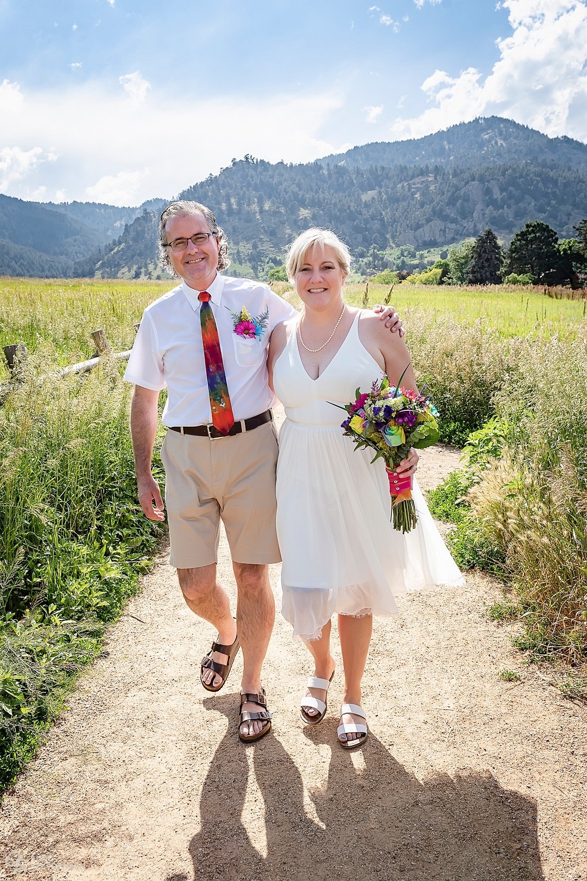 The bride and groom in white with tie-dye accessories and brightly colored bouquet walk along a pathway at Chautauqua Park, Boulder.