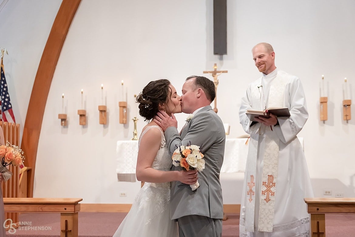 A newly married couple kiss during their Lutheran church ceremony as the officiant looks on and smiles.