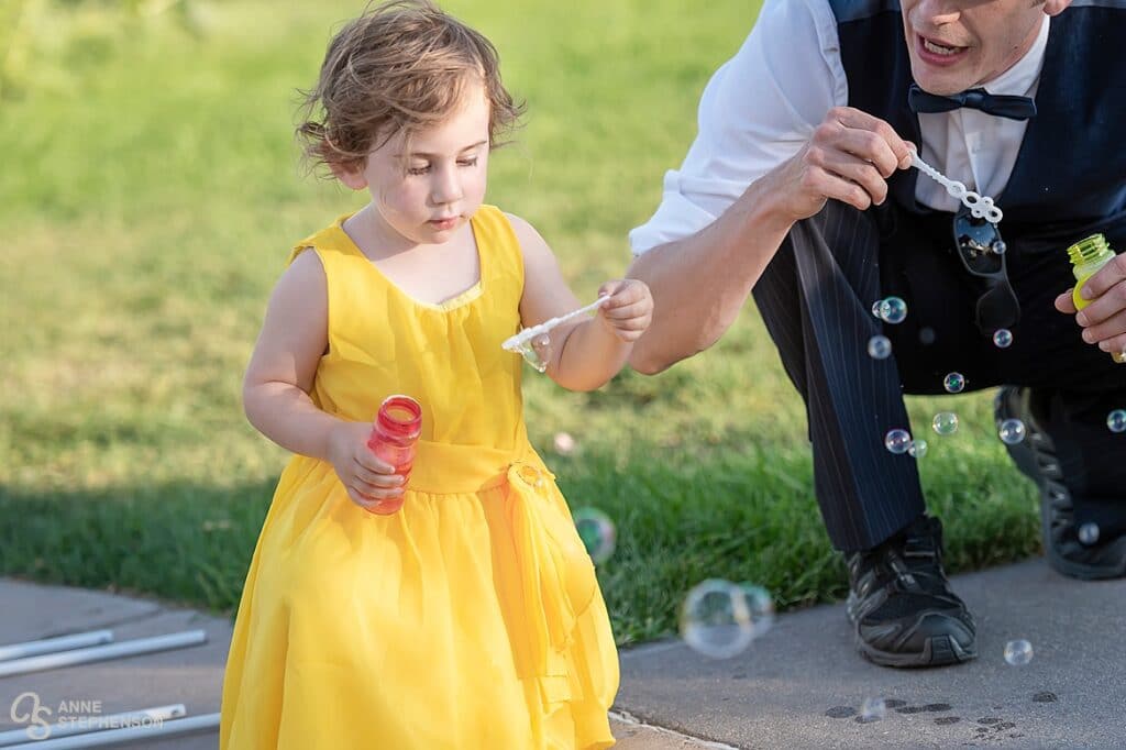 A flower girl in a yellow dress blows bubbles.
