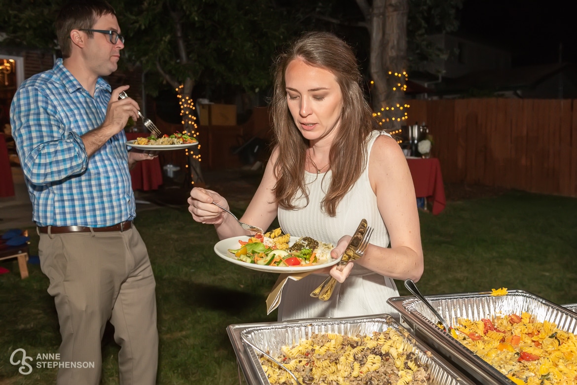 The bride at a backyard wedding celebration fills her plate with pasta salad from a delectable food buffet created by her friend.