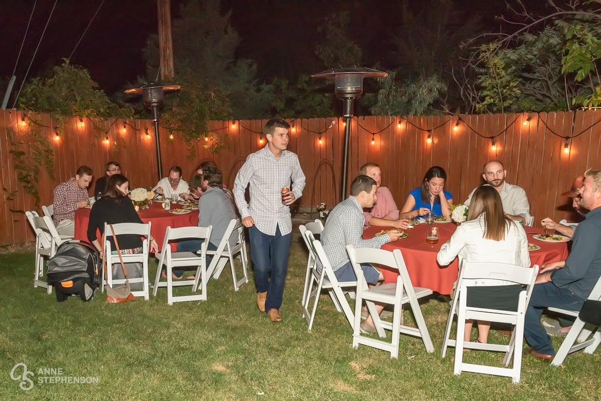 Guests assemble at tables with red cloths and sit down on white chairs at this casual evening backyard wedding party.