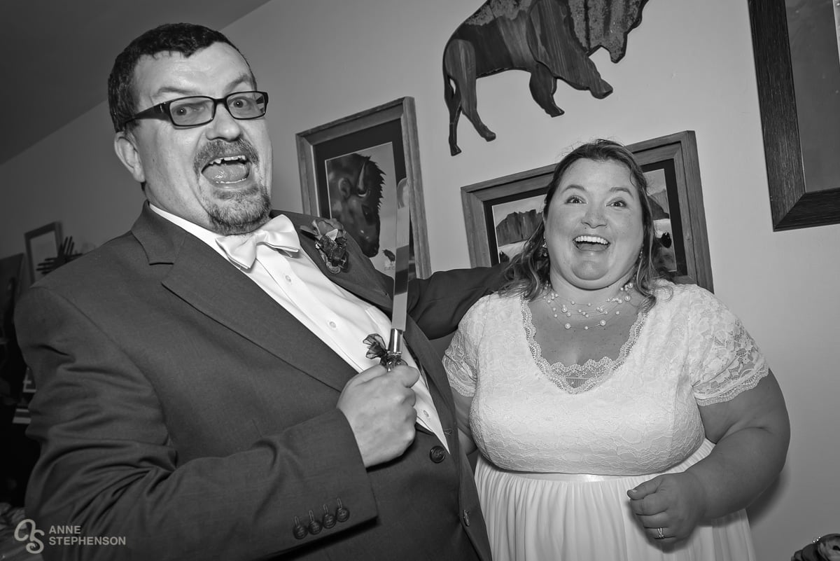 A black and white photo of the groom holding the knife in a horror movie style and placing his arm around the bride.