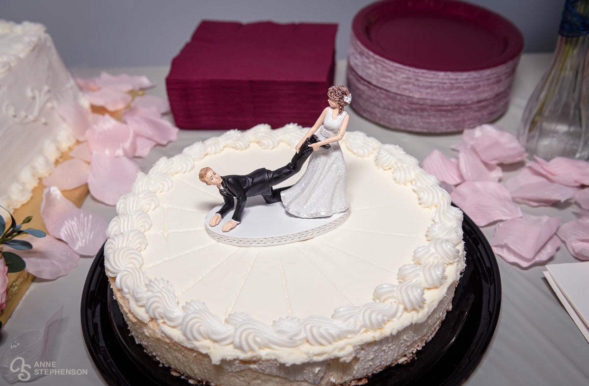 A grooms cake made of cream cheese with a funny cake topper of the bride pulling the groom's leg as he scrambles on the ground to escape.