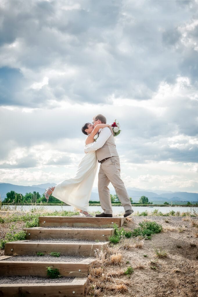A woman kicks up her leg and kisses her husband just after their marriage. Behind them are beautiful clouds and a mountain view.