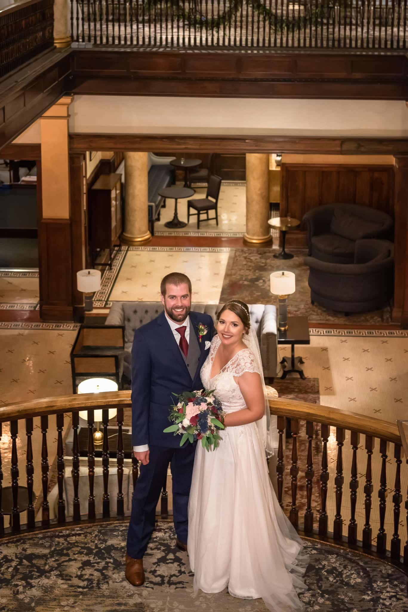 A bride and groom stand midway down the stairs on the landing of the Hotel Boulderado with the lobby in the background.