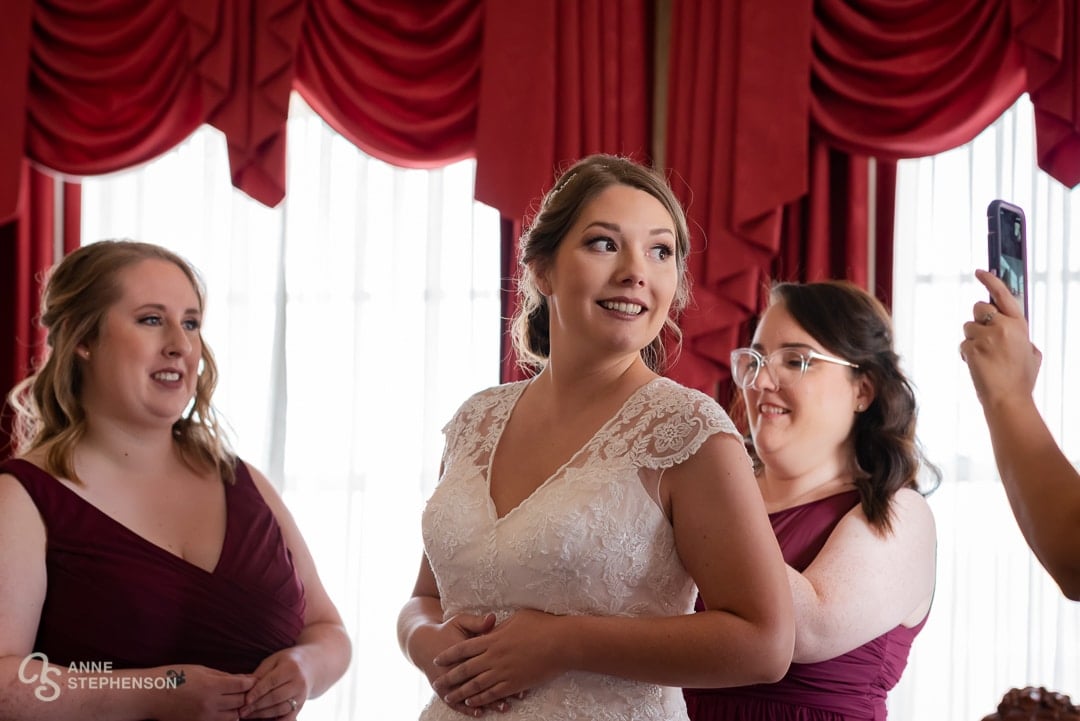 In the getting ready room with dark red curtains, a bride turns to a cell phone and smiles before  talking to a relative who was unable to make the wedding.