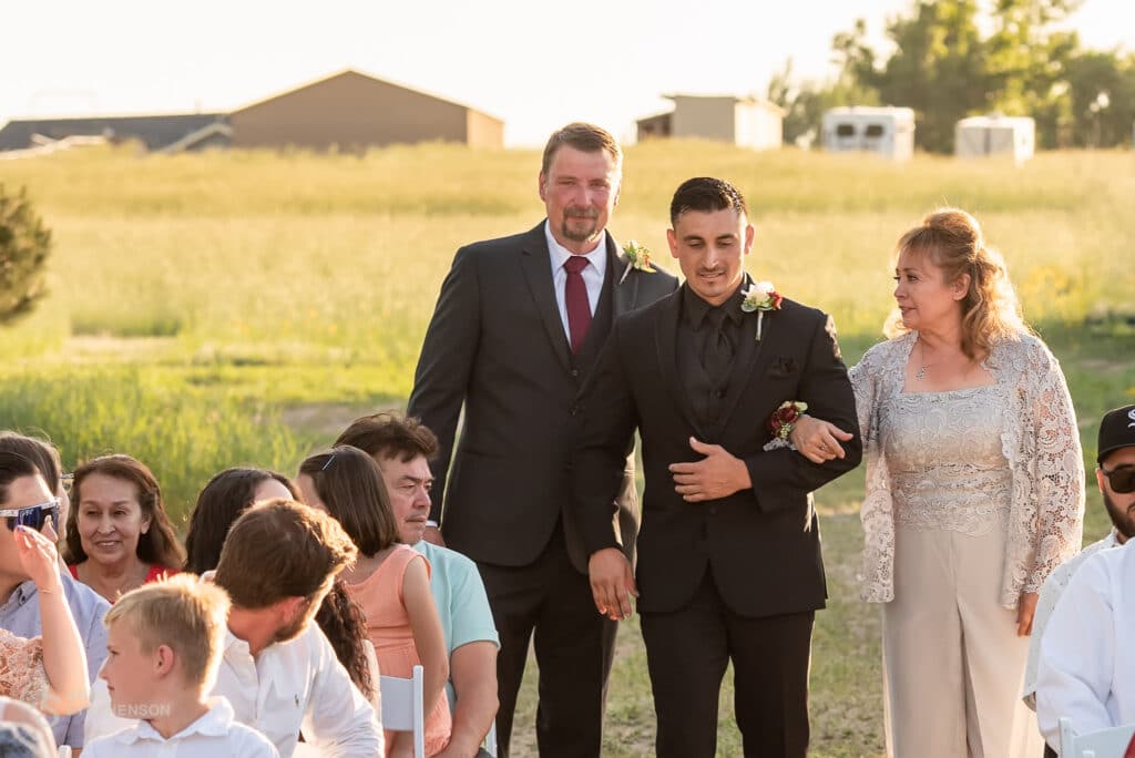 The groom walks down the aisle, escorted by his dad on the left and mom on the right.