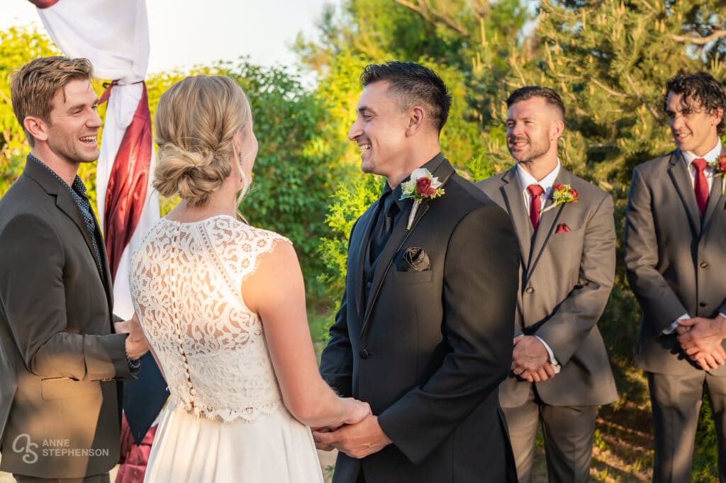 The groom turns to the officiant and laughs while holding hands with his bride.