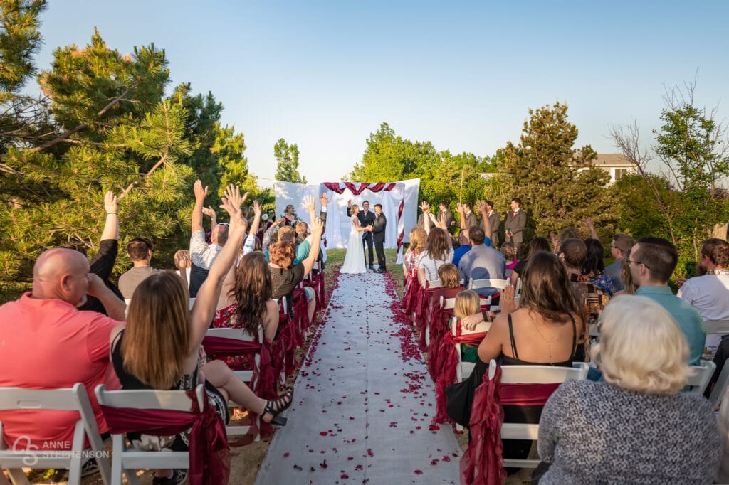 Wide view of the wedding ceremony from the last row showing the guests raising their hands in agreement that they are happy the wedding is taking place.