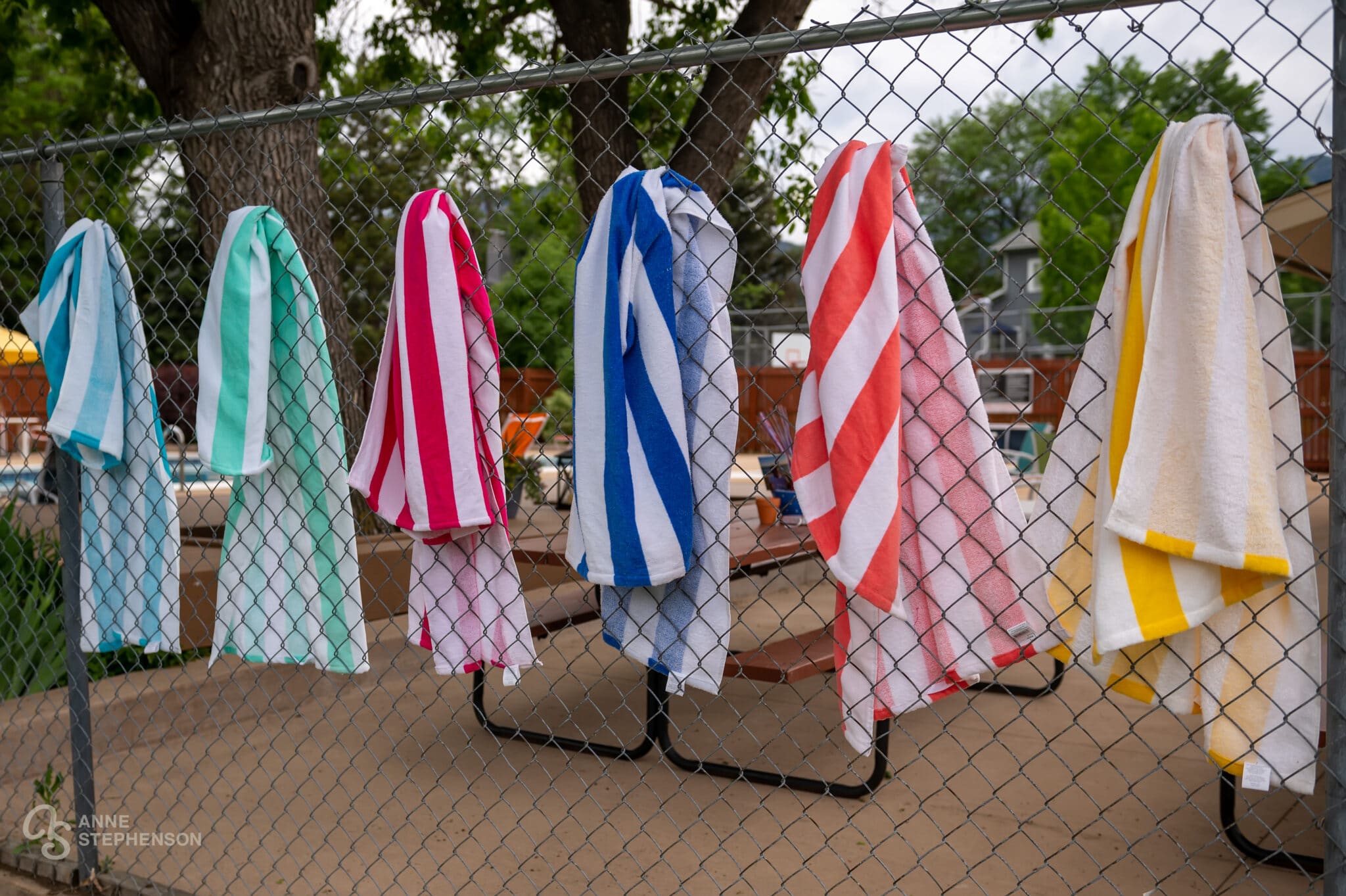 A row of different colored striped beach towels hanging on a chain link fence outside of a pool.