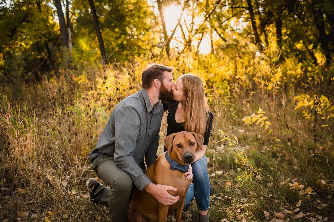 An engaged couple in an autumn wooded area kisses as they hold their dog close.