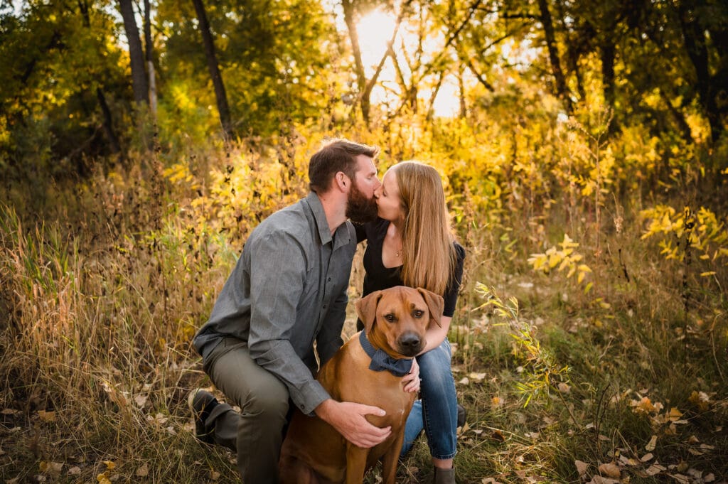 An engaged couple kisses during their autumn photo session as their dog looks on.
