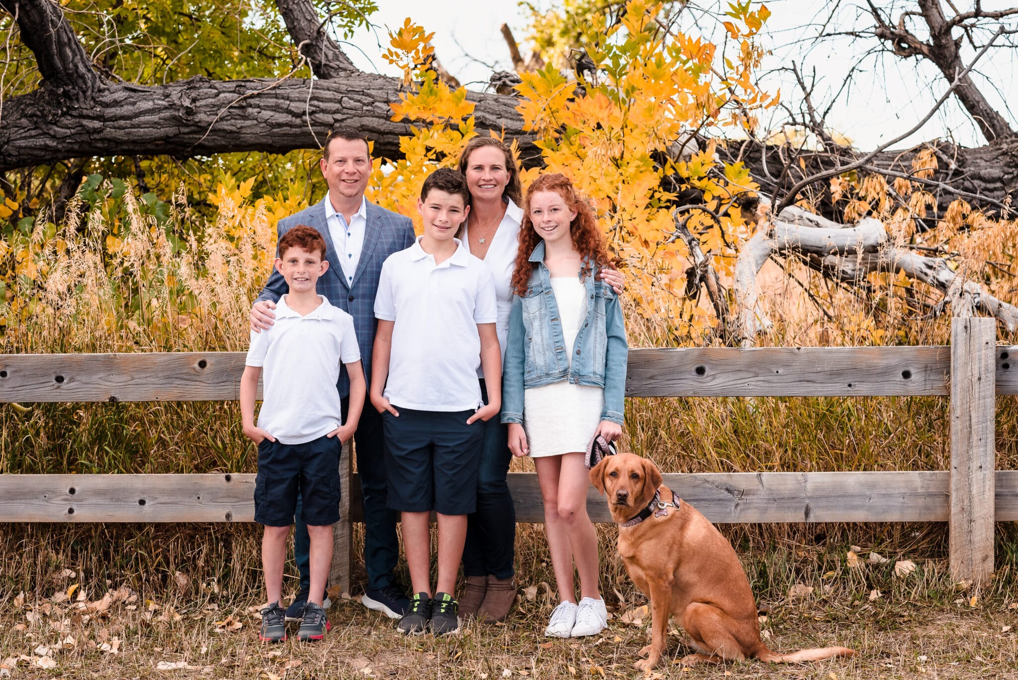 A family of five--mom, dad, two boys and a daughter--lean against a fence as their dog looks on during their autumn family portrait session.