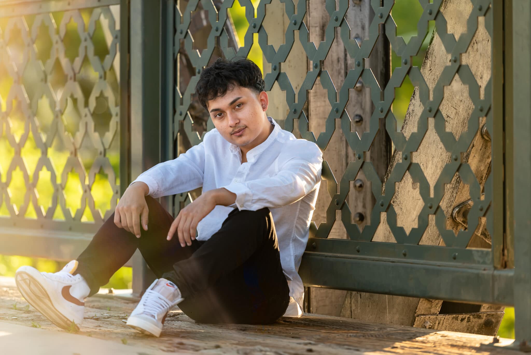 A high school senior sits on the ground resting his back against a green metal lattice work bridge.