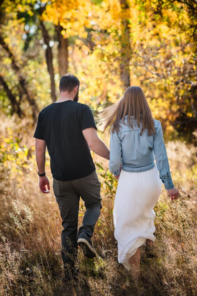 An engaged couple hold hands and walk thru the golden woods in autumn.