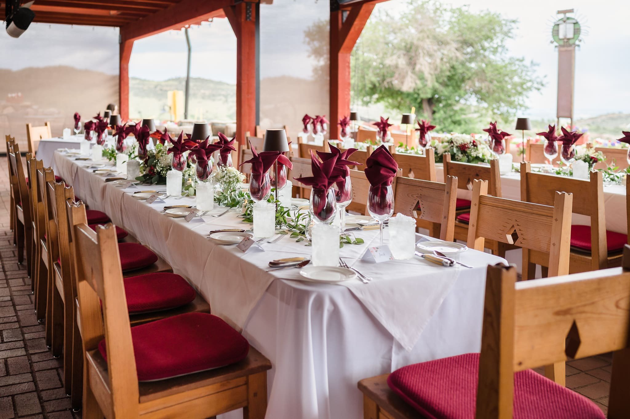 A long banquet table set up with wine glasses, small plates, silverware, and red napkins underneath the overhang on the patio at The Fort in Morrison, Colorado.