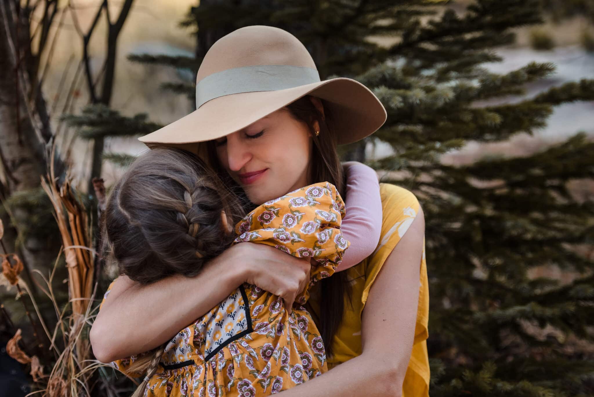 A mother wearing a wide brimmed hat closes her eyes and smiles as she holds her young daughter tightly for a hug during a family portrait session.