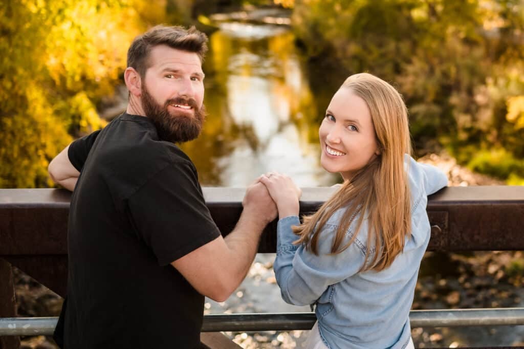 An engaged couple on a bridge with fall colors in the background.