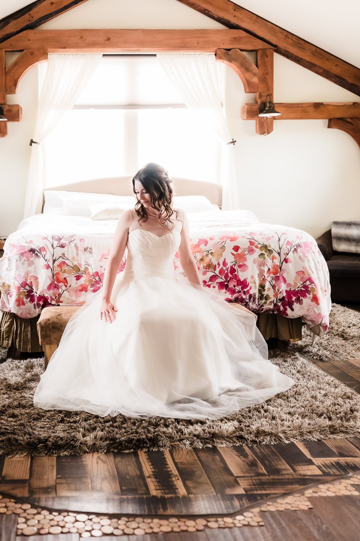 The bride sits on a bench in front of the bed with beautiful window light behind her in the getting ready room at 1C Barn, Monument, Colorado.