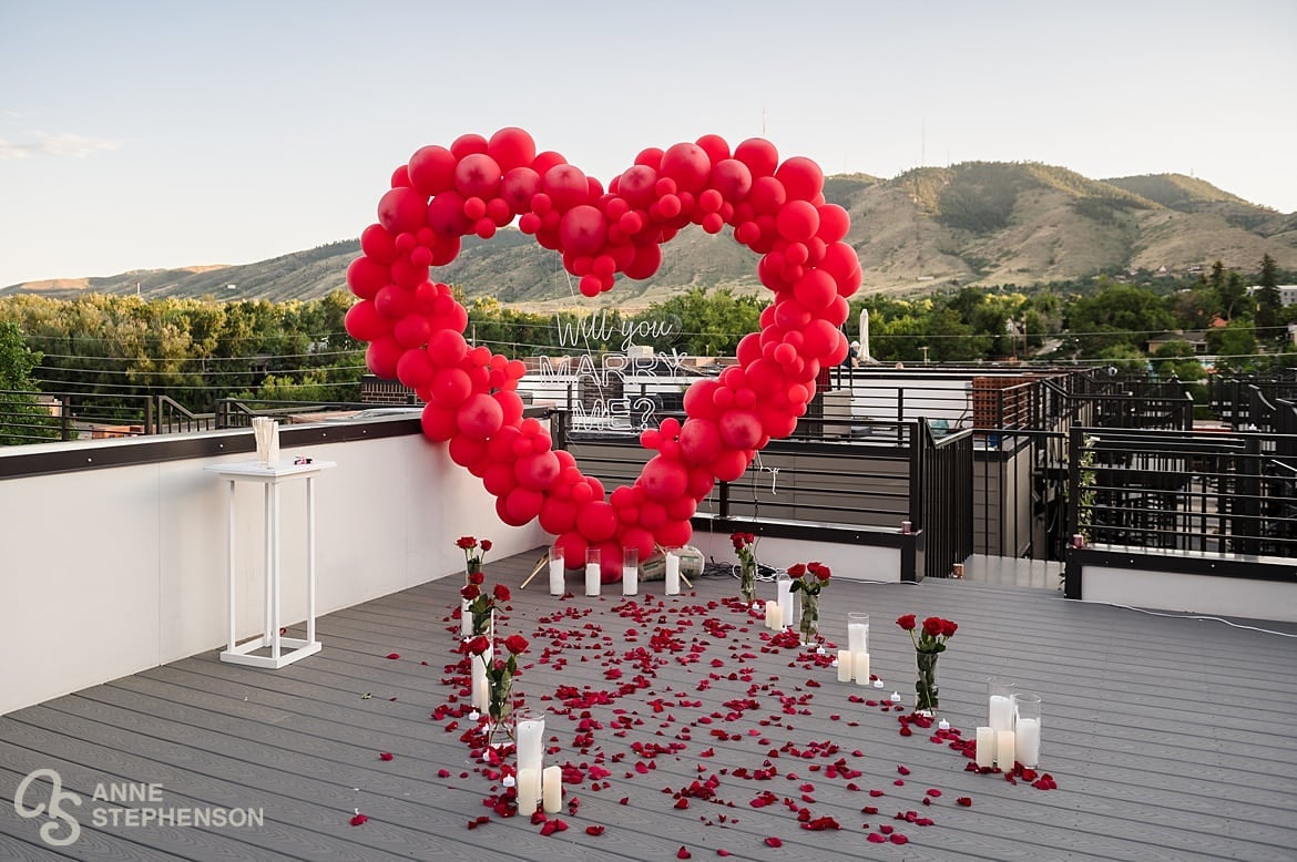 A romantic proposal scene complete with red balloons, red roses, white candles, and strewn red rose petals.