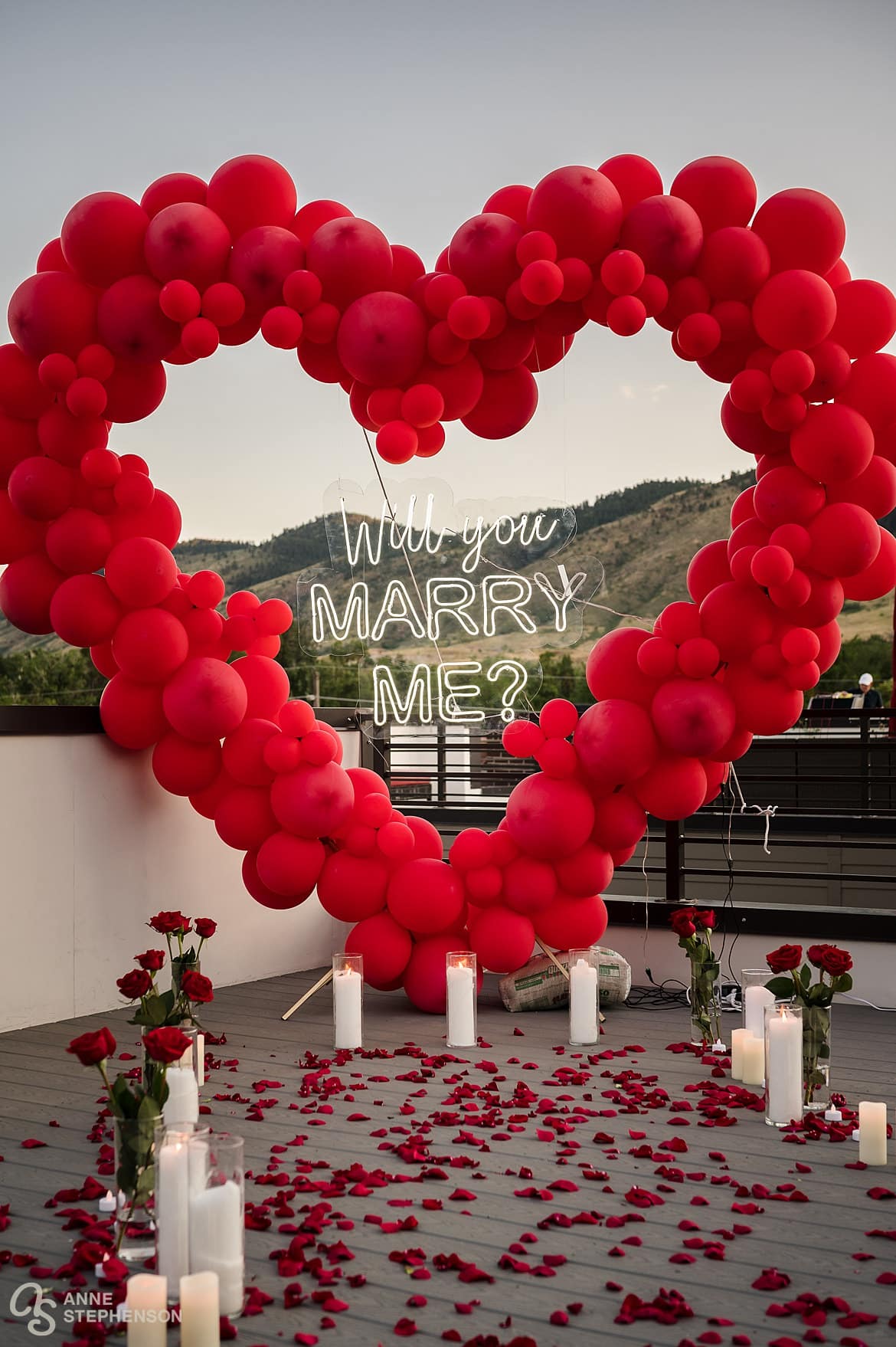 A romantic outdoor proposal scene in the summer golden hour with a heart-shaped red balloon display, and rose-strewn path lined with red roses and candles.