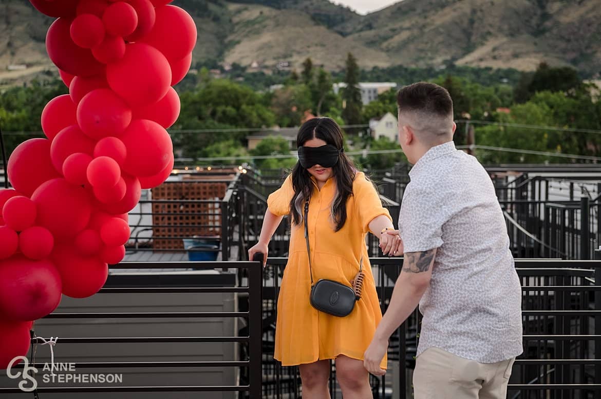 Best engagement photos on a rooftop in Golden, Colorado start with a boyfriend leading his blindfolded girlfriend to a decorated scene.