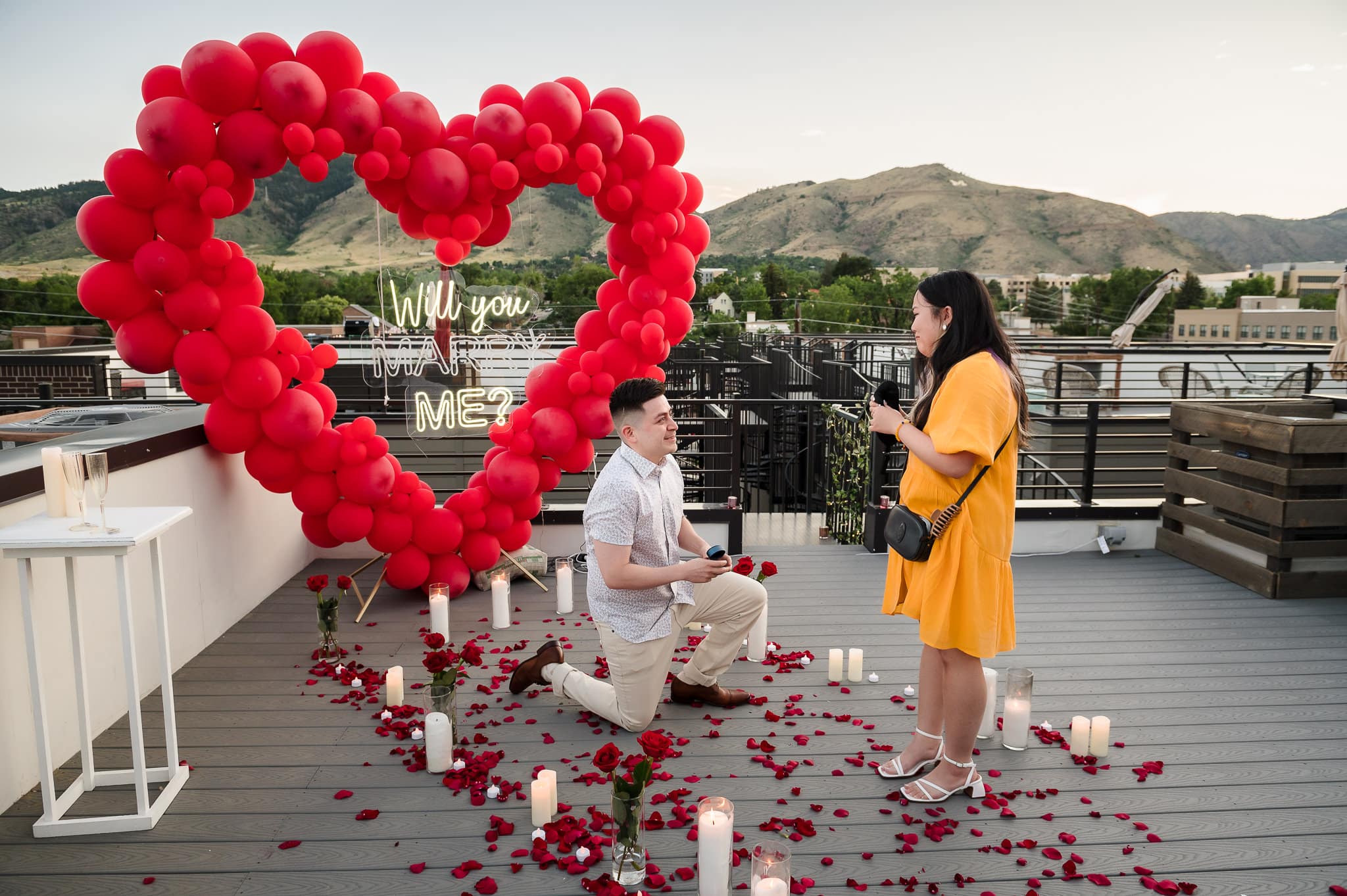 A young man on a rooftop balcony at sunset bends his knee in front of a balloon shaped heart that says "marry me" amid strewn rose petals and proposes to his girlfriend.